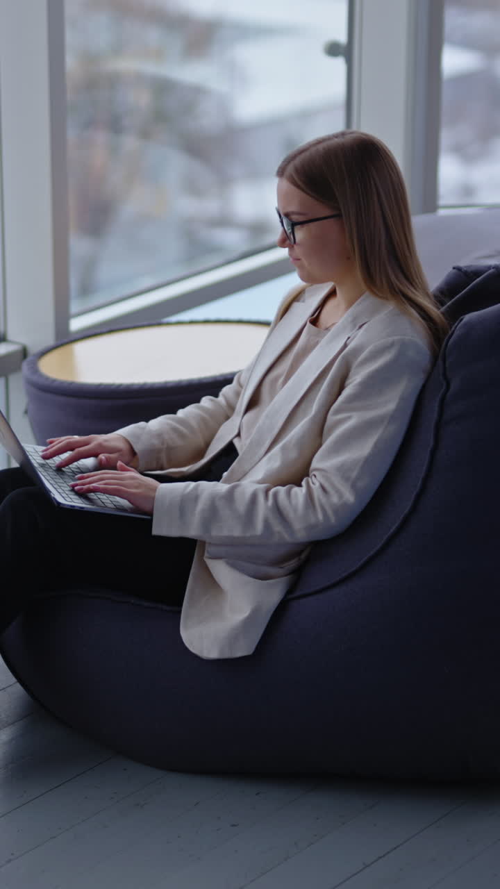 Young lady works at laptop sitting in the bean bag chair. Woman is tired a little and yawns covering her mouth. Side view. Vertical video