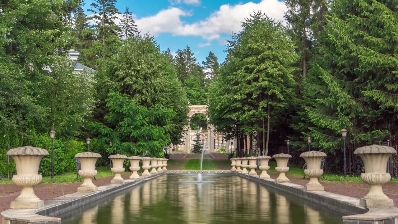 夏の公園または森の緑の木々と茂みのタイムラプス。湖とアーチのある噴水がある小道。空には雲。