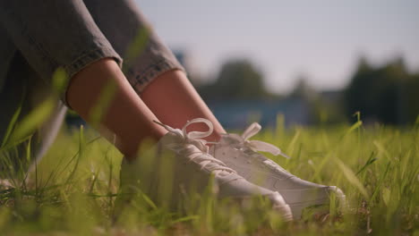 close-up of woman s legs in jeans and white canvas shoes, surrounded by green grass gently swaying in breeze, with sunlight softly illuminating her shoes, distant blurred background