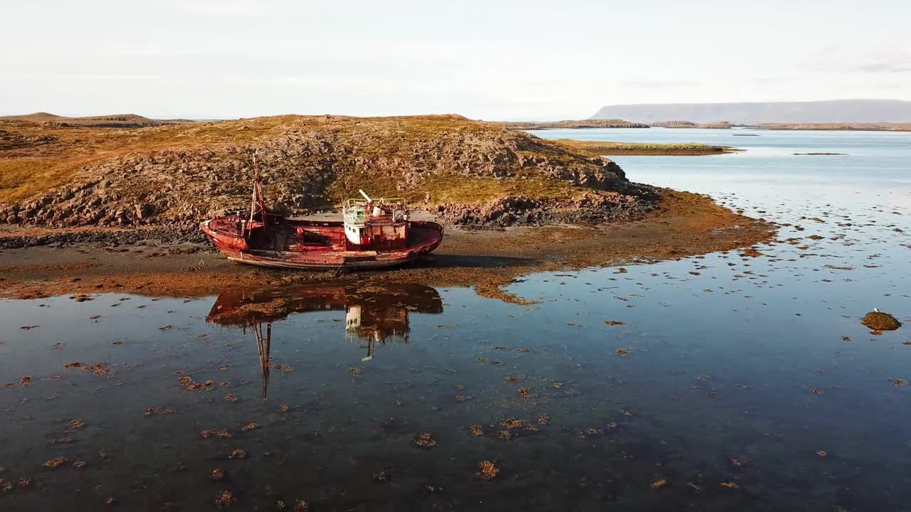 Aerial view capturing the Breiðabólsstaður boat graveyard at sunset, with abandoned, decaying boats on a reddish beach, surrounded by tranquil waters and small islands, drone pulling out