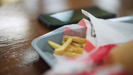 person placing phone beside container of fries on smooth wooden table, closeup of snack setup with warm light reflecting on surface, casual break moment capturing texture and simple lifestyle