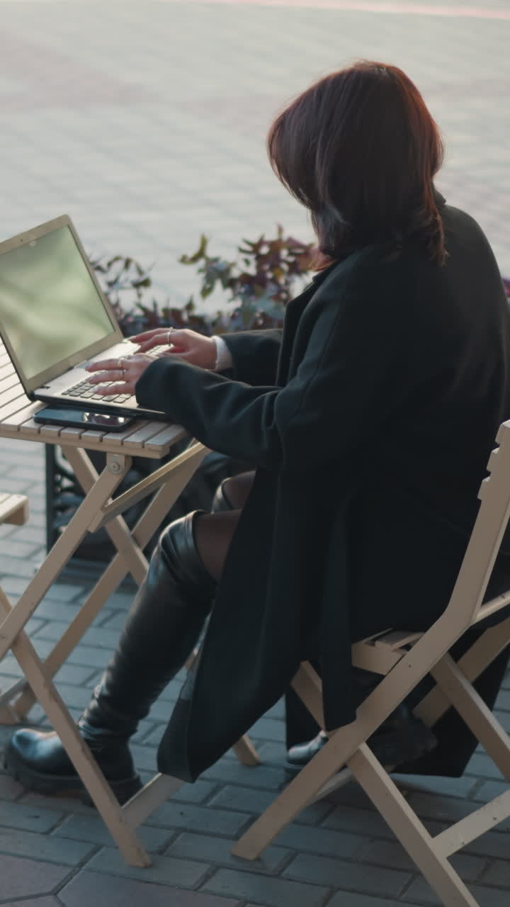 카페에서 야외에서 노트북으로 일하는 프리랜서, 나무 의자에 둘러싸인 식물과 테이블에 앉아, 뒷면에 포장 된 산책로와 건물이 흐릿하게 배경에 배치 된 드백