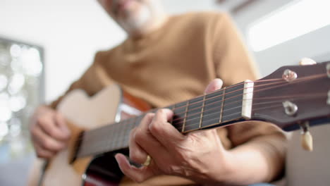 midsection of senior biracial man sitting playing guitar at home, selective focus, slow motion