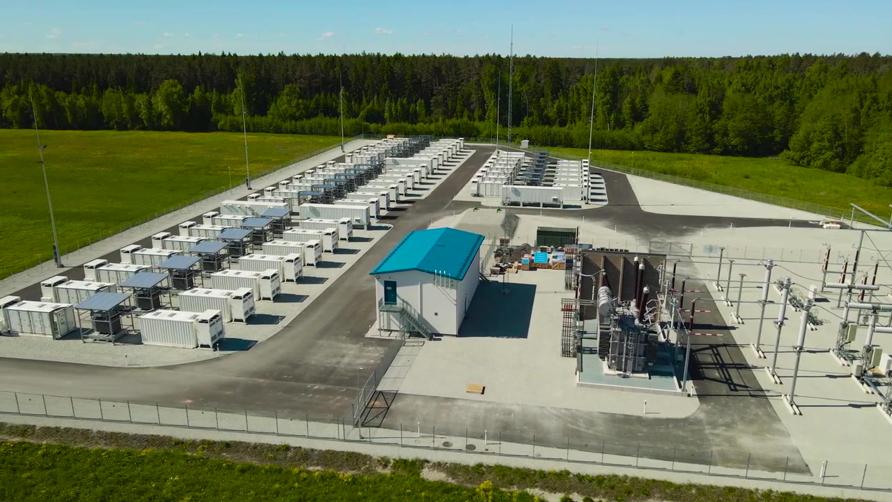 Aerial view circling a battery energy storage system plant with its large capacity containers for storing renewable power, located in a rural green field with a dense forest in the background
