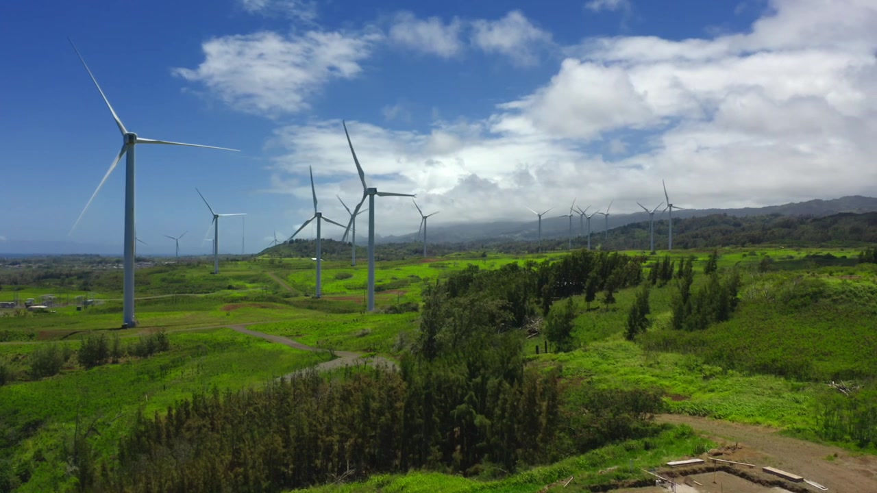 Dolly aéreo hacia aerogeneradores girando con elegancia en vastos campos verdes de Kahuku en la costa norte de Oahu, energía renovable en un entorno natural y tranquilo