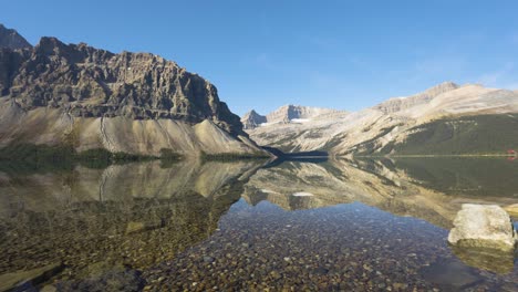 Breite-Handgehaltene-Schwenkaufnahme-Von-Kristallklarem-Seewasser-Mit-Bergen-Und-Wald-An-Einem-Sonnigen-Tag