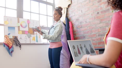 Diverse-female-fashion-designers-using-laptop-and-tablet-discussing-designs-at-studio,-slow-motion