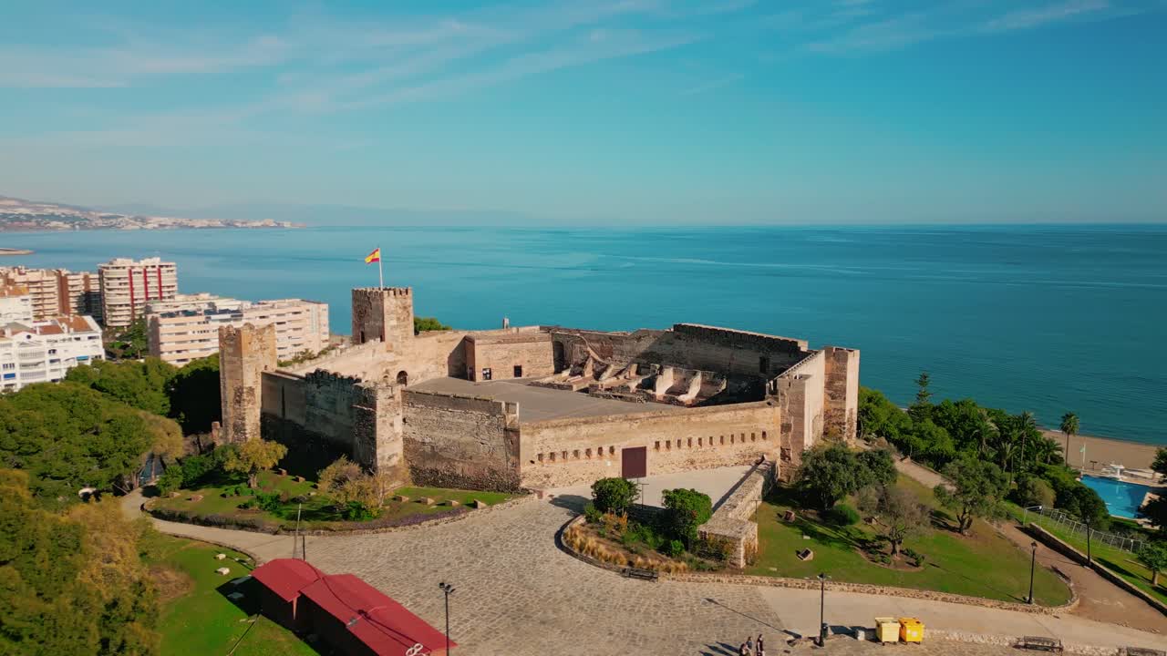 Órbita aérea do Castelo de Sohail em Fuengirola, Costa do Sol, Espanha, com a bandeira espanhola tremulando sobre a fortaleza histórica enquanto a vista se estende do horizonte da cidade em direção ao horizonte do Mediterrâneo.