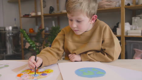 blond kid painting a earth on a paper sitting at a table in a craft workshop