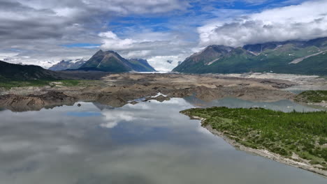 stunning overcast sky reflects in the smooth surface of the lake. drone rising slowly over the waterscape revealing the view on the gorgeous mountains. alaska wilderness