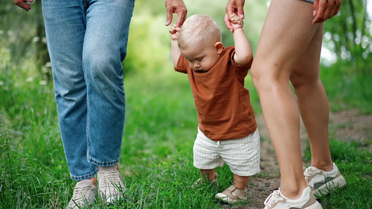 Parents hold their baby by the hands walking in the park. Cute toddler boy learning to walk outdoors.