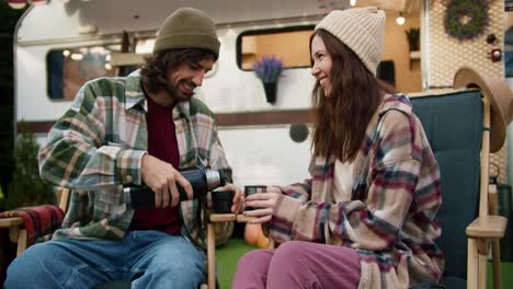 A-happy-brunette-guy-in-a-green-hat-and-a-plaid-shirt-pours-tea-from-a-thermos-to-his-brunette-girlfriend-in-a-hat-and-They-drink-tea-while-sitting-on-green-chairs-near-a-trailer-during-a-picnic-outside-the-city-in-the-summer