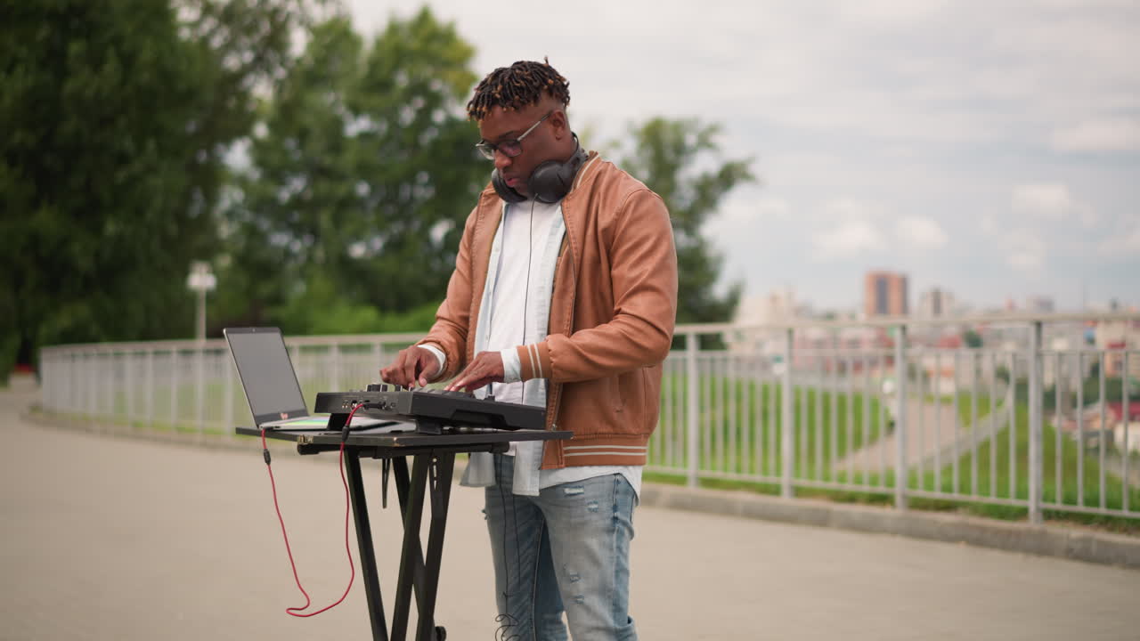 Parkta stant üzerinde dizüstü bilgisayar ve MIDI kontrolcü ile açık havada klavye seti performansı sergileyen siyahi DJ, kahverengi ceketli, kulaklıklı, şehir silueti ve korkuluk arka planında, gündüz atmosferinde, enerjik miksaj ve sokak sanatçısı enerjisiyle.