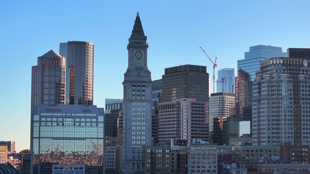 Custom House Tower stands among Boston Financial District skyscrapers. Glass facades glimmer around historic clock tower. Gliding overhead city skyline, aerial.