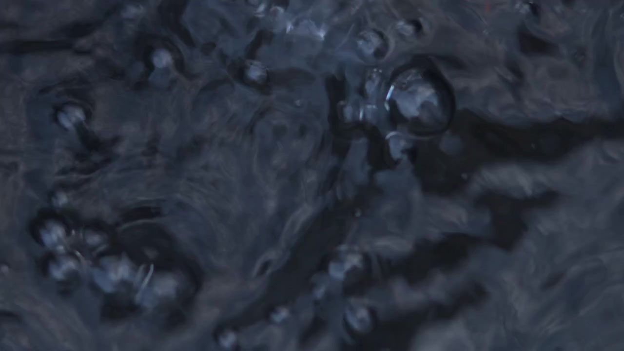 Primo piano di una superficie liquida scura con bolle attive, schiuma vorticosa e vapore che sale sotto una luce rossa calda, creando una texture industriale astratta e suggestiva.