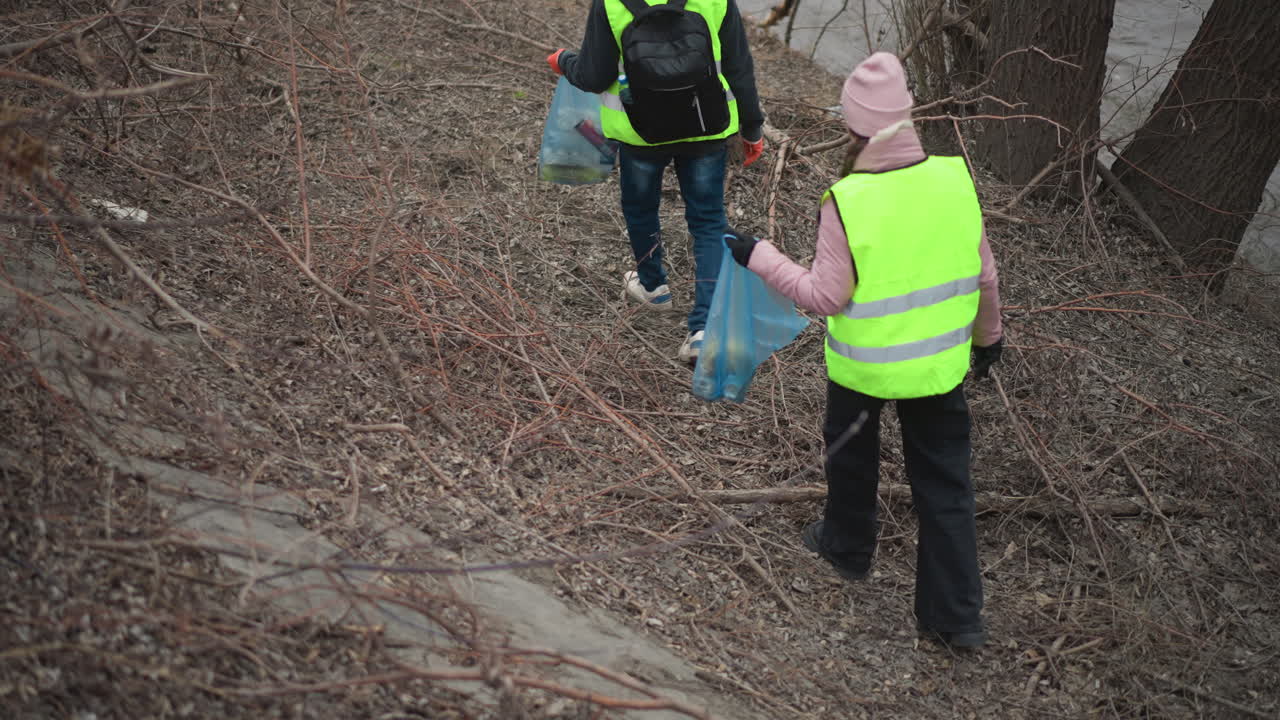 Volunteers in reflective safety vests walking along riverbank collecting litter in blue plastic bags during environmental cleanup, wearing gloves and warm clothing to protect nature