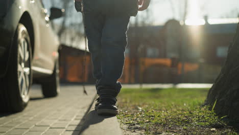 a close-up shot of a child walking along a pavement holding a stick, missed his step before climbing back onto the pavement the child is dressed in a shiny black jacket, surrounded by