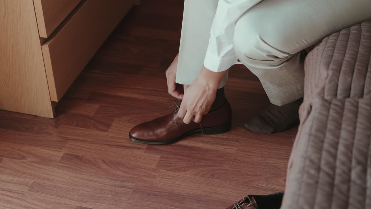 Close up of a man tying brown leather shoes while sitting on a bed indoors
