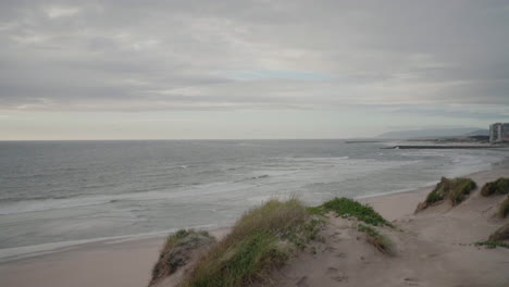 Overcast-coastal-view-with-sandy-dunes,-ocean-waves,-and-distant-buildings