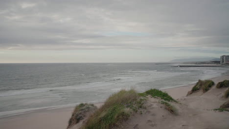 Overcast coastal view with sandy dunes, ocean waves, and distant buildings