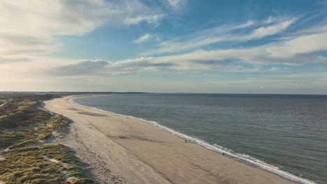 Toma-Aérea-Larga-Y-Alta-De-Un-Mar-En-Calma,-Playa-Casi-Vacía-Y-Caminos-A-Través-De-Dunas-Cubiertas-De-Hierba-Al-Atardecer-En-Un-Hermoso-Día