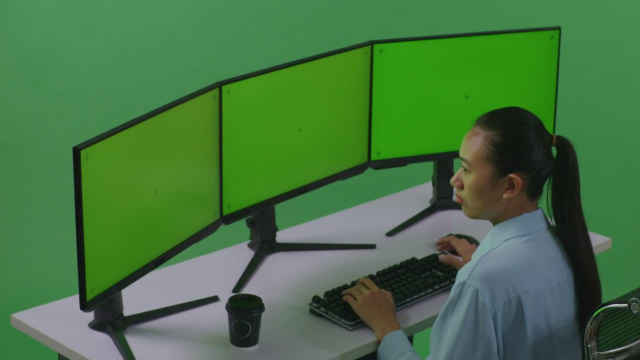 vista laterale di una donna asiatica che lavora con diversi monitor di computer in uno studio a schermo verde
