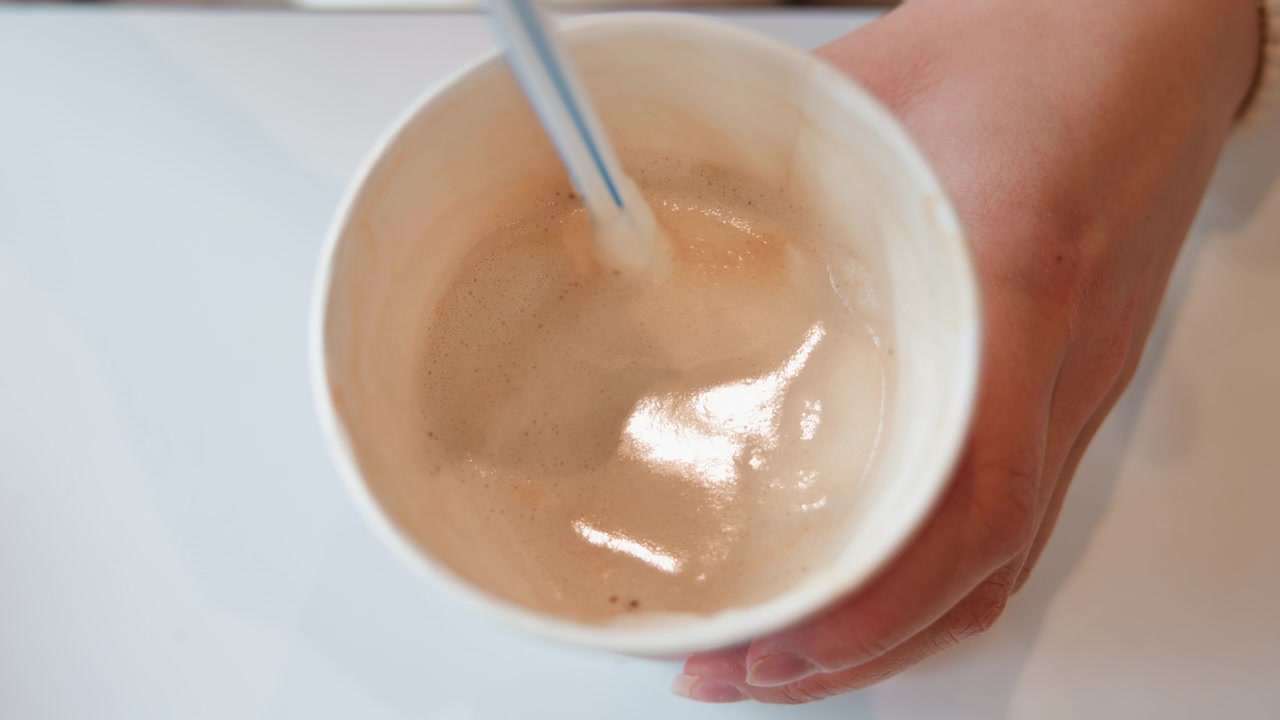Vista ravvicinata dall'alto di mani che tengono una tazza di ceramica bianca mentre un cucchiaino mescola caffè o schiuma di latte, creando un vortice visibile, un residuo lucido e un leggero movimento durante la preparazione di una bevanda mattutina informale.