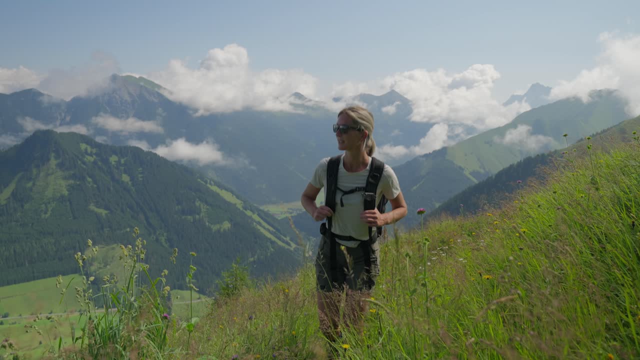 A woman hikes up a grassy mountain trail surrounded by scenic views and distant clouds