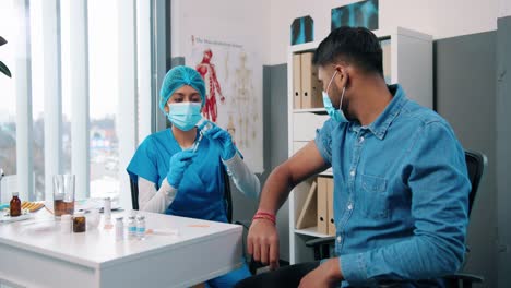 portrait of hindu beautiful young woman nurse or doctor virologist injecting handsome male patient sitting in hospital cabinet in medical mask, healthcare concept, coronavirus vaccination, covid 19