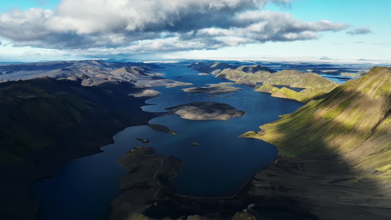 Bredt luftfoto over en lang vulkansk innsjø omgitt av robuste islandske fjell, med drivende skygger som avslører lagdelte rygger, øyer og dypt blått vann som strekker seg inn i horisonten