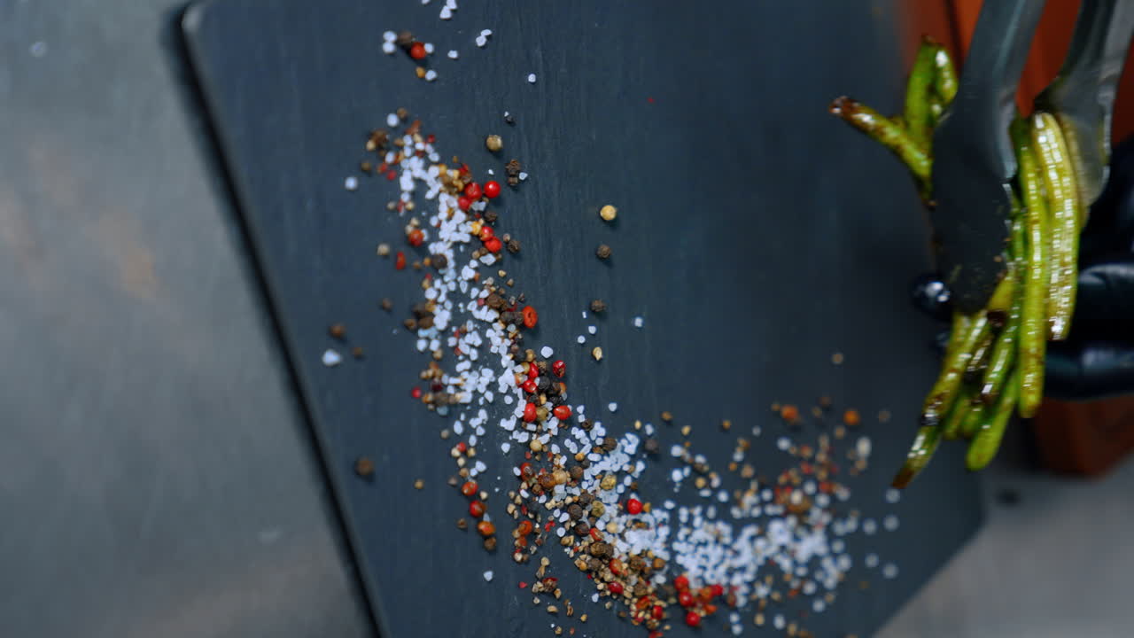 Black charcuterie board with salt and peppers on. Hands in black gloves use tongs to lay the fried beans on the board. Vertical screen. Close up.