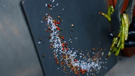 black charcuterie board with salt and peppers on. hands in black gloves use tongs to lay the fried beans on the board. vertical screen. close up.
