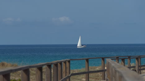 Pequeño-Velero-Blanco-Navegando-Rápido-En-El-Mar-Azul-Cerca-De-La-Costa,-Cielo-Despejado-Con-Pocas-Nubes,-Barreras-De-Un-Bonito-Camino-De-Madera-En-Primer-Plano