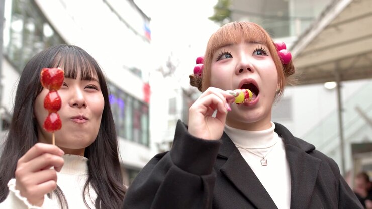 Due donne asiatiche gustano snack tanghulu alla fragola e all'uva all'aperto in un vivace ambiente urbano.