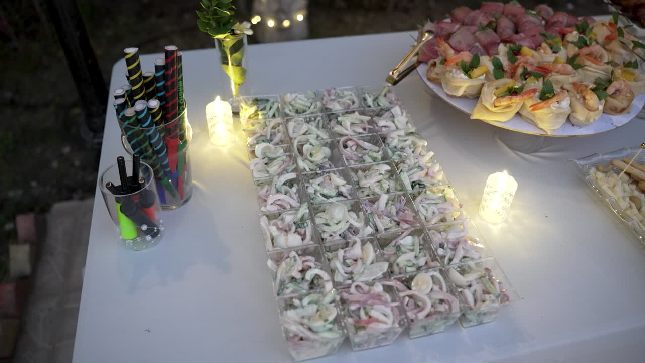 Guests enjoy an outdoor gathering featuring a variety of tasty appetizers arranged neatly on tables. The setting is illuminated gently with soft lights, creating a warm atmosphere
