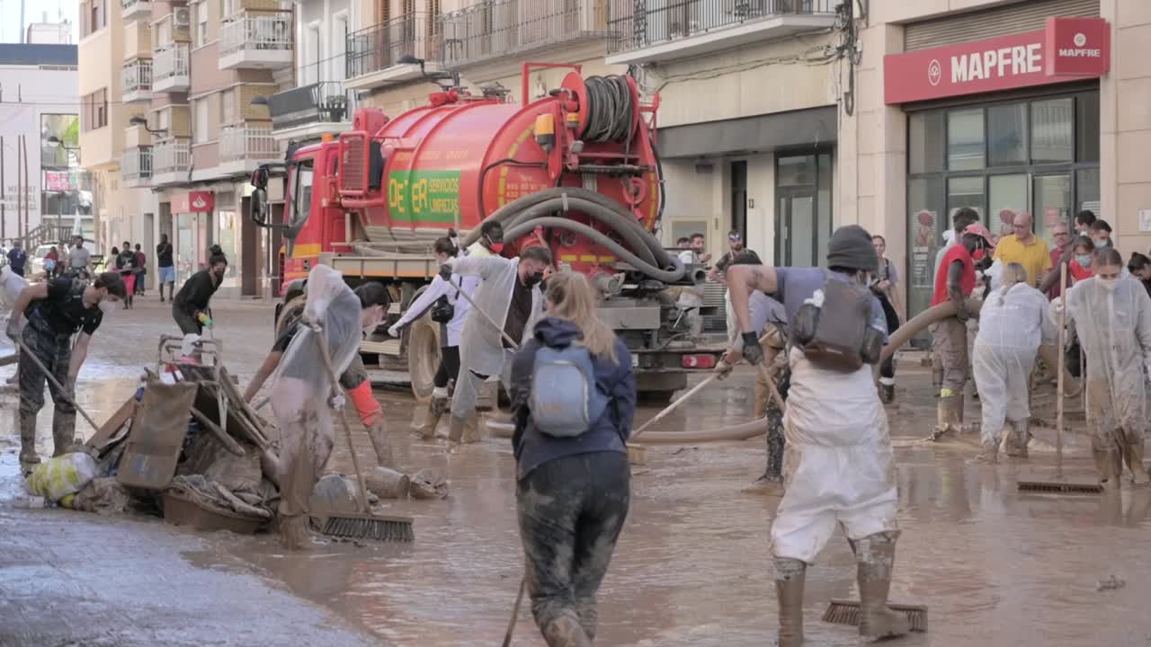Volunteers work together to clean a muddy street after a flood in an urban area with a cleaning and debris removal truck in Valencia. People use brooms and shovels to clear debris and water