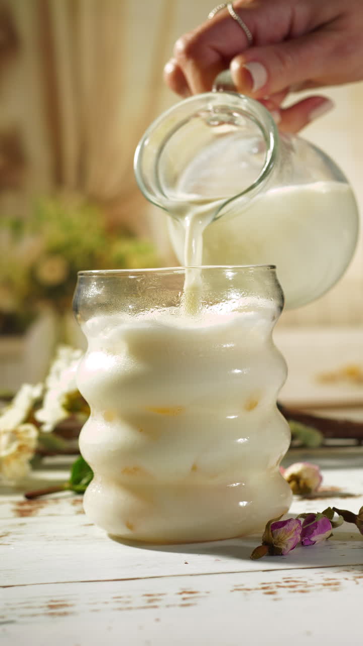 A hand pours creamy vegan oat milk or cow milk from a clear glass pitcher into a bubble-shaped glass on a rustic white wooden table adorned with dried rose blossoms, bathed in warm morning sunlight.
