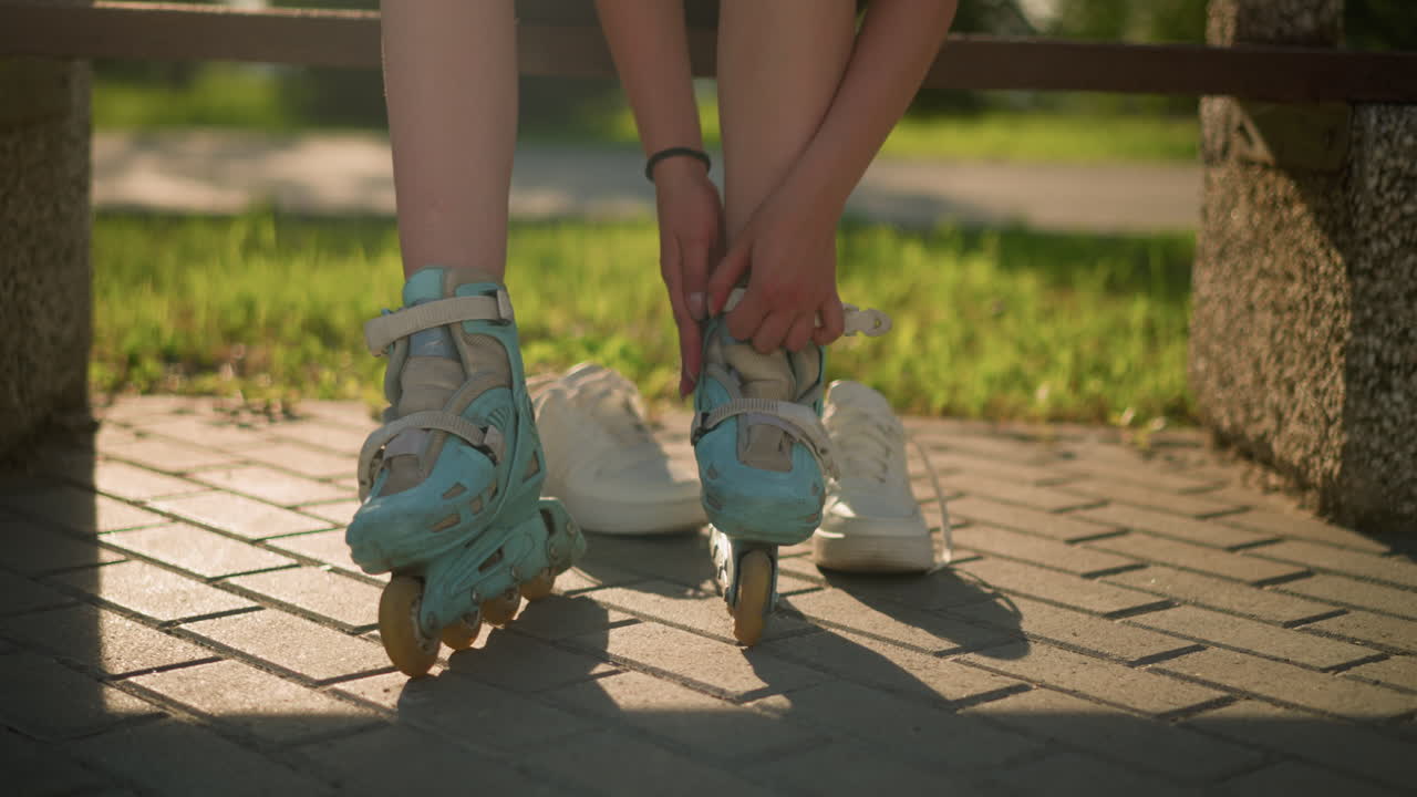Leg view of individual sitting on bench adjusting strap of cyan roller skates with black bangle on right hand, while white sneakers rest behind on pavement