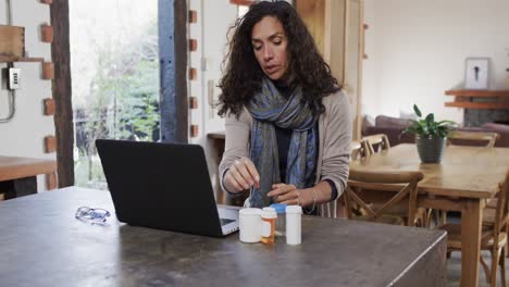 Biracial-woman-with-cold-holding-medication-and-making-video-call-using-laptop-at-home,-slow-motion