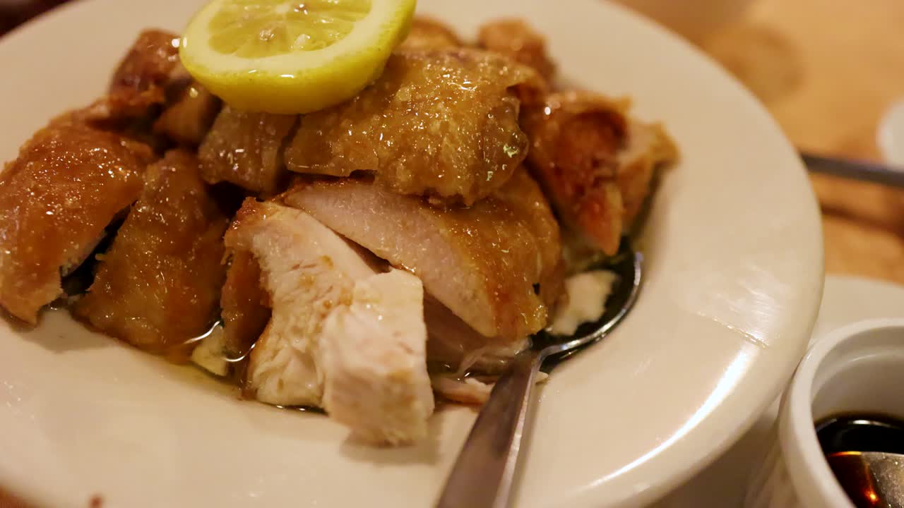 Close-up of lemon crispy chicken on a white plate in a warmly lit Chinese restaurant, with a spoon and dipping sauce visible, camera slowly panning