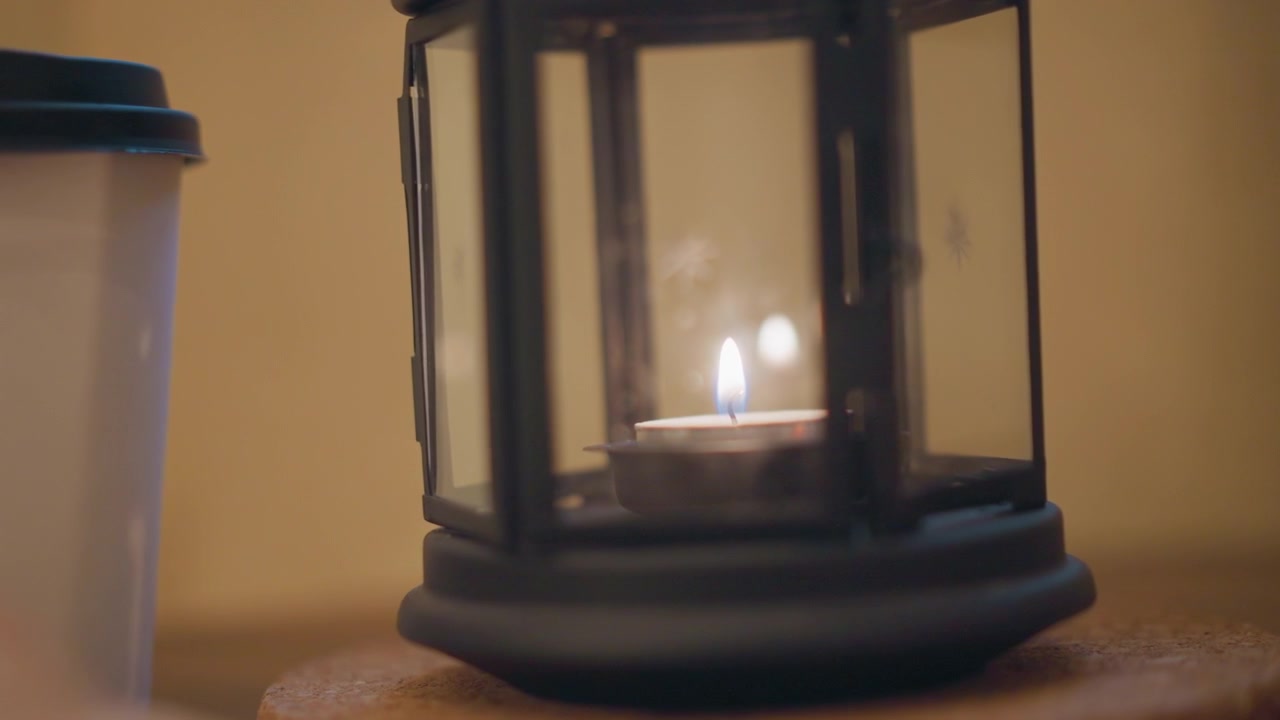 Primo piano di una candela accesa in una lanterna di vetro accanto a una tazza bianca con coperchio nero su una superficie di legno. La fiamma della candela tremola dolcemente, proiettando una luce calda e ombre tenui intorno agli oggetti su uno sfondo neutro.