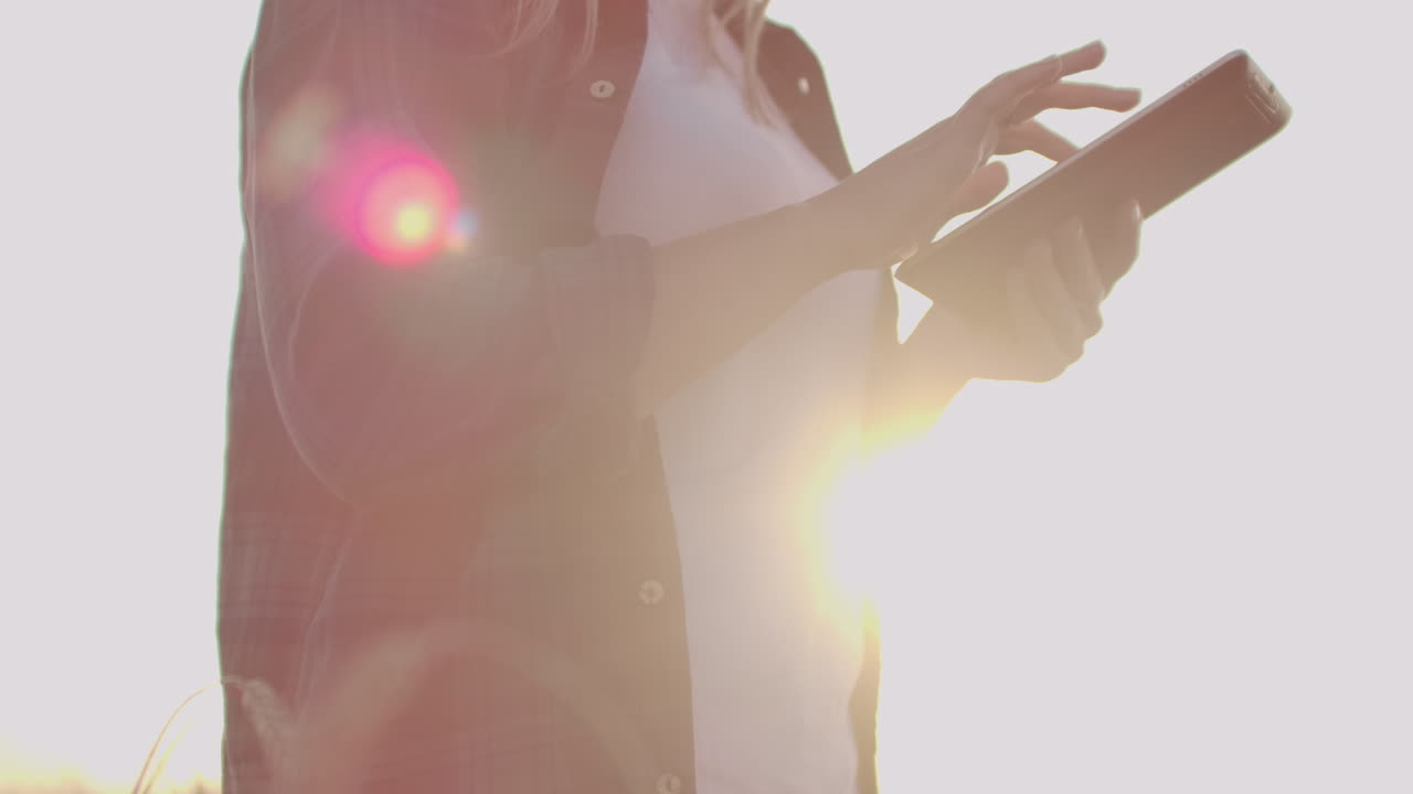 손에 태블릿 컴퓨터를 들고 칠색 셔츠를 입은 여성 농부가 해가 지는 동안 밀을 가로질러 걸어가고 있습니다.