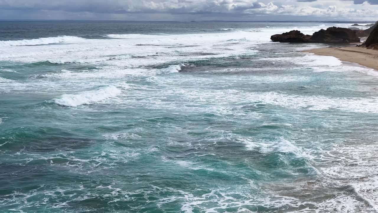 Dynamic aerial footage of turquoise waves breaking on sandy shore under cloudy daylight at Portsea