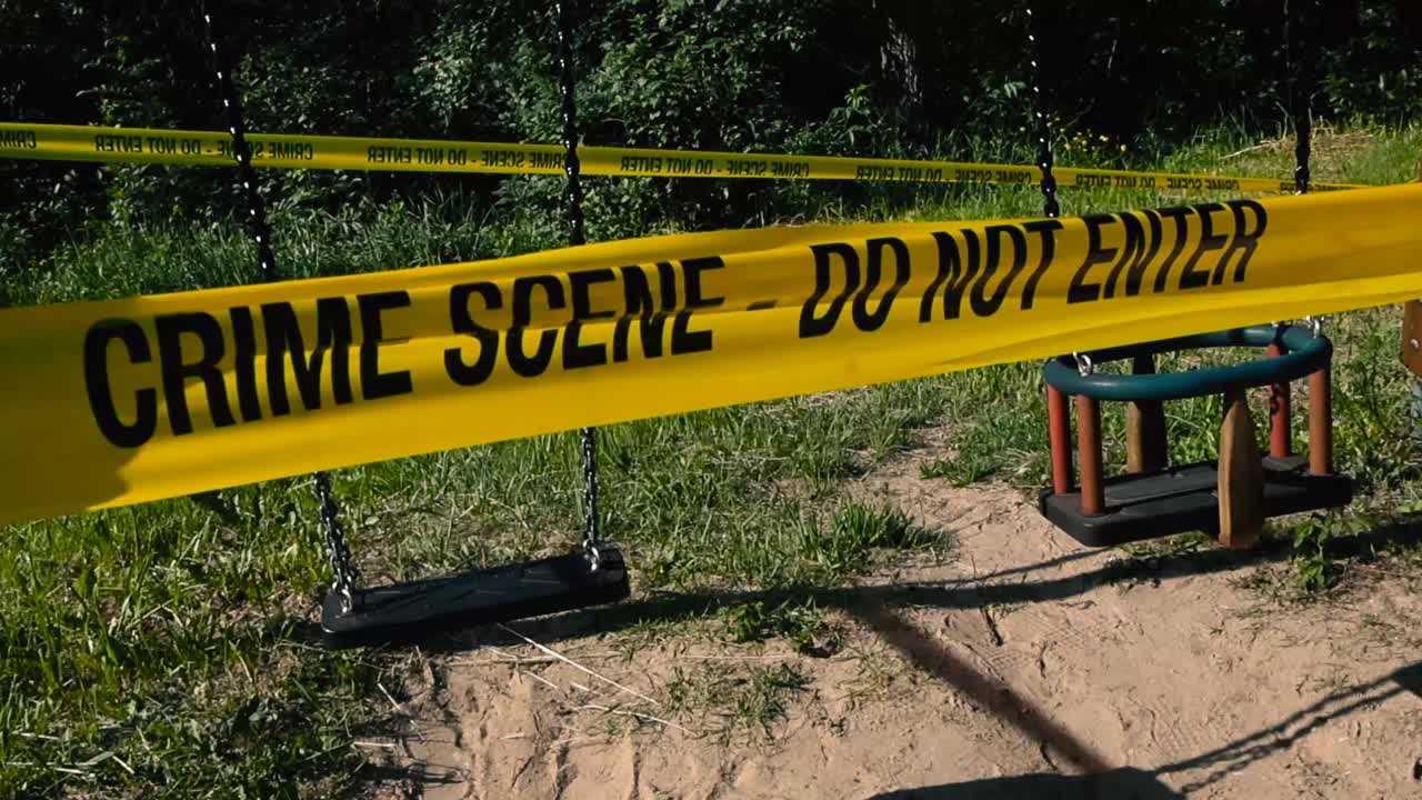 Yellow and vibrant colored Crime Scene Do Not Enter investigation ribbon tape put in front and around two enpty swings swinging at a playground during a sunny summer day. Sandy ground, police tape.