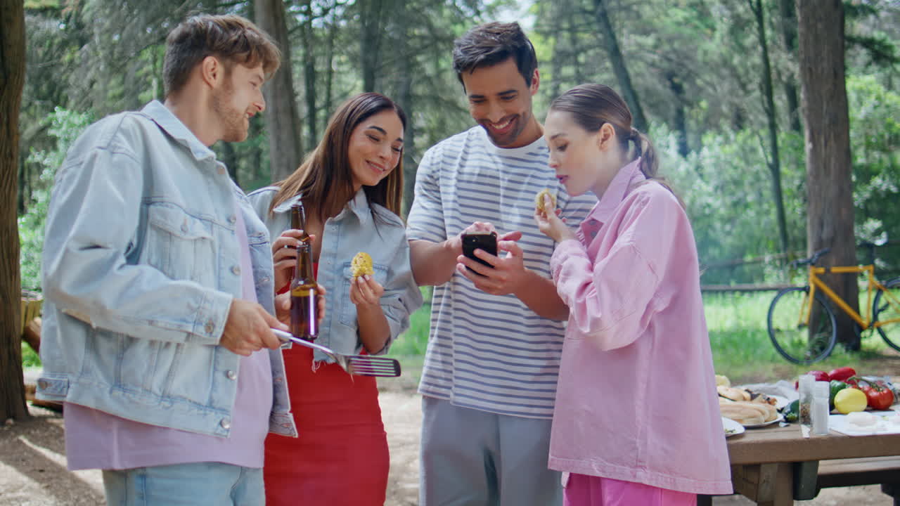 Group friends bonding barbecue at forest picnic. Smiling man showing smartphone