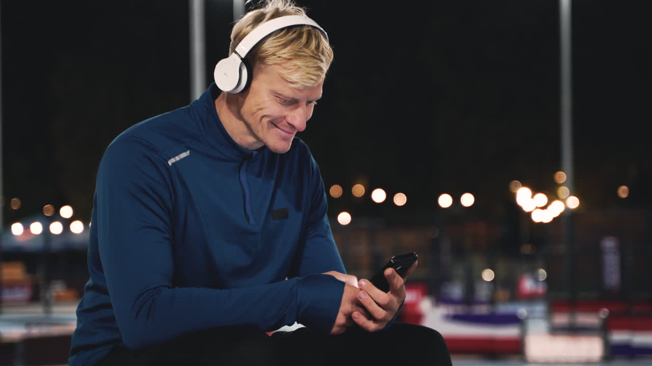Smiling Sportsman Sitting At Park Listening Music With Bluetooth Headphones, Texting On His Mobile Phone And Looking Around Him While Taking A Break During His Training Session At Night