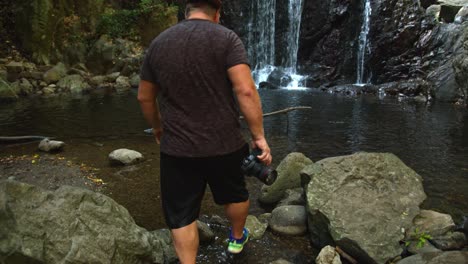 Young-strong-man-walking-towards-waterfall-with-camera-and-aims-for-photo