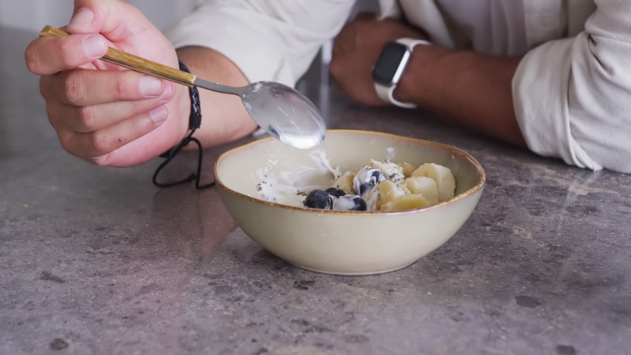 African American adult male seeing bowl reaching with spoon and stirring yogurt to eat at counter