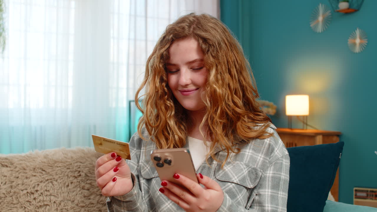 Young woman sitting on sofa holding bank card and smartphone making online payment or money transfer