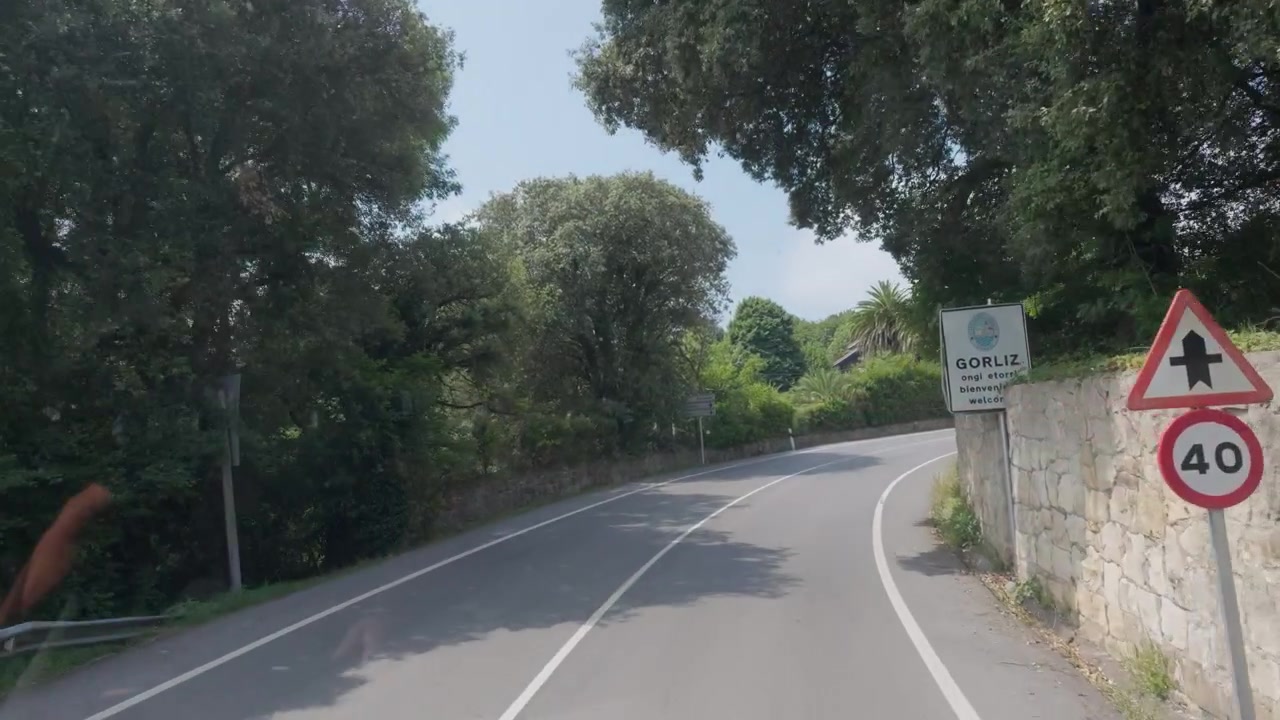 Strada di campagna che curva tra alberi verdi, passando un muro di pietra e segnali stradali, suggerendo viaggio e percorso
